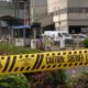 Yellow caution tape reading 'DO NOT CROSS' and 'CAUTION: SHERIFF LINE' in foreground at a restricted area. Industrial buildings and vehicles in background.