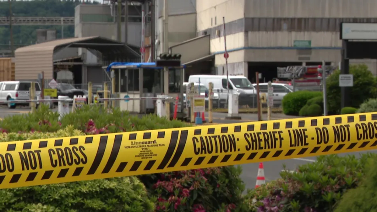 Yellow caution tape reading 'DO NOT CROSS' and 'CAUTION: SHERIFF LINE' in foreground at a restricted area. Industrial buildings and vehicles in background.