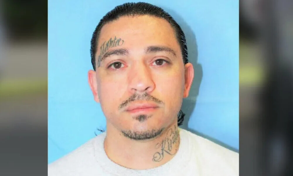 Mugshot of a man with a tattoo on his left temple and neck, neutral expression, blue background.