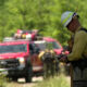 Firefighter in turnout gear with helmet checks a radio as red fire trucks respond on a forested rural road.