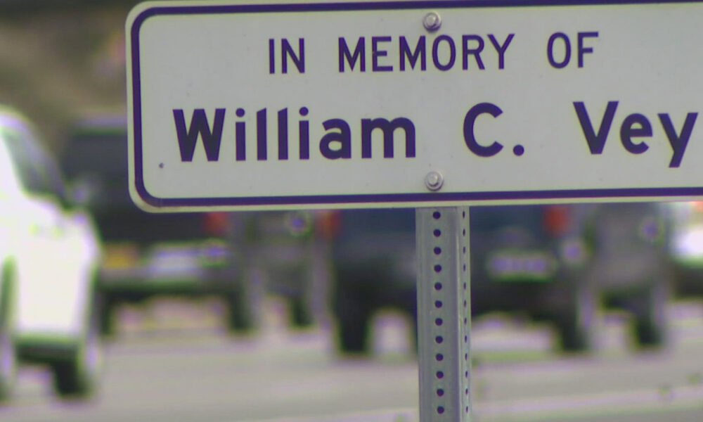 Memorial sign reading "In Memory Of William C. Vey" on a metal post with a blurred street scene in the background.
