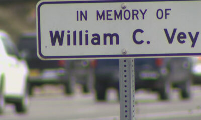 Memorial sign reading "In Memory Of William C. Vey" on a metal post with a blurred street scene in the background.
