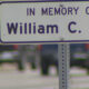Memorial sign reading "In Memory Of William C. Vey" on a metal post with a blurred street scene in the background.