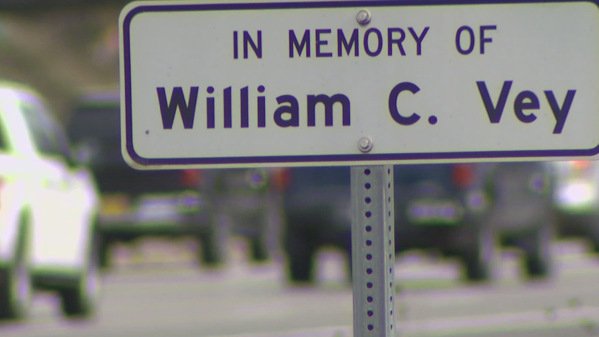Memorial sign reading "In Memory Of William C. Vey" on a metal post with a blurred street scene in the background.