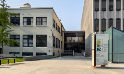Campus plaza with a beige two-story building on the left, a tall concrete building on the right, a bench in the foreground, and a campus map sign nearby.
