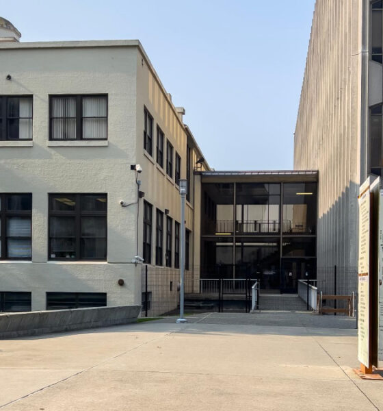 Campus plaza with a beige two-story building on the left, a tall concrete building on the right, a bench in the foreground, and a campus map sign nearby.