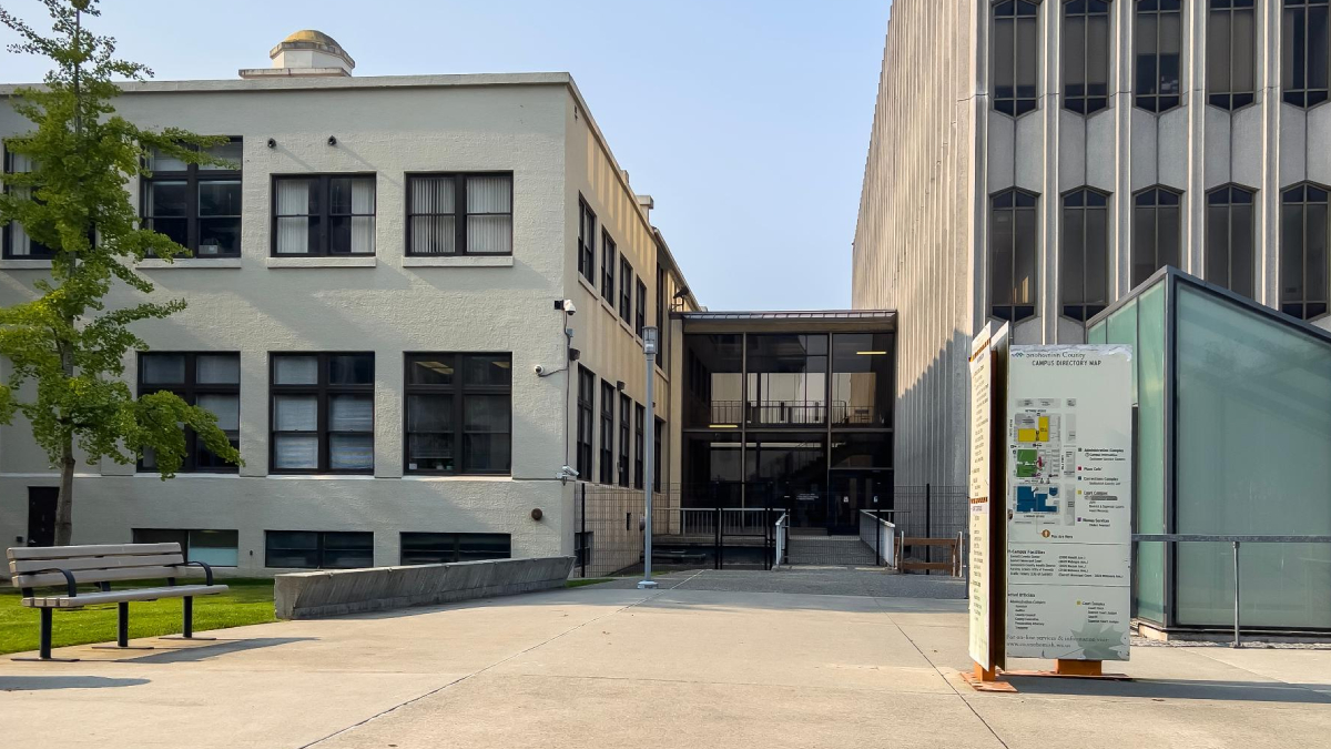 Campus plaza with a beige two-story building on the left, a tall concrete building on the right, a bench in the foreground, and a campus map sign nearby.