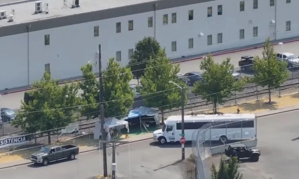 A white tour bus parked near a chain-link fence with small tents and trees in a fenced lot behind a large gray building.