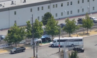 A white tour bus parked near a chain-link fence with small tents and trees in a fenced lot behind a large gray building.