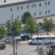 A white tour bus parked near a chain-link fence with small tents and trees in a fenced lot behind a large gray building.