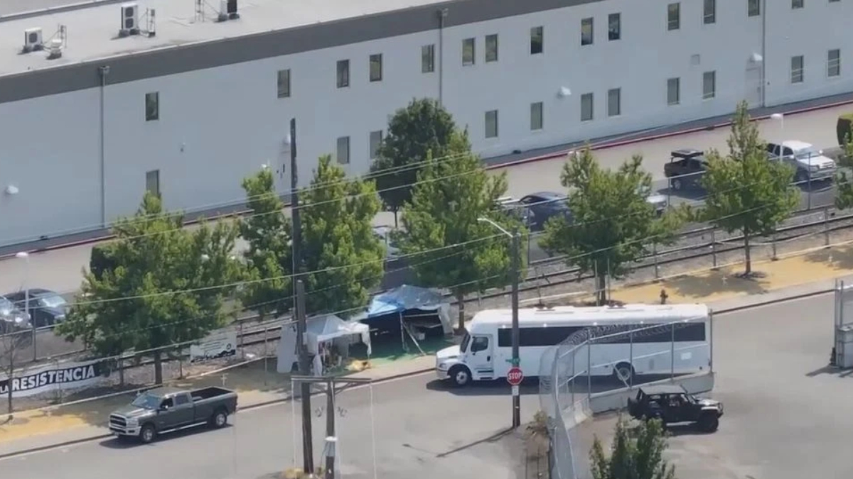 A white tour bus parked near a chain-link fence with small tents and trees in a fenced lot behind a large gray building.