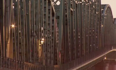 Long metal arch bridge with vertical suspender cables, illuminated streetlights along the deck at dusk.