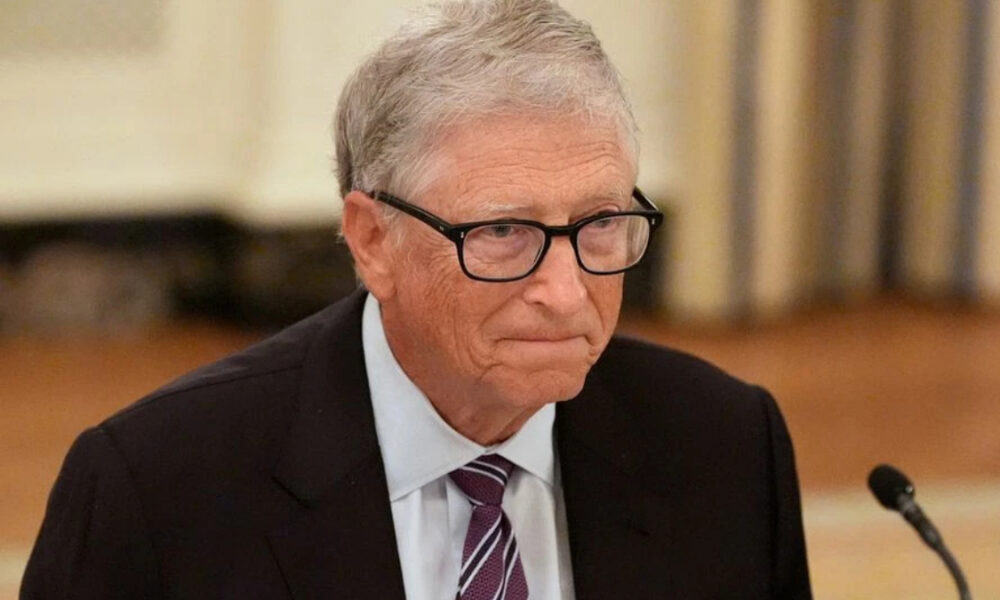 Older man with gray hair and glasses in a dark suit and striped tie at a conference table with a microphone nearby.