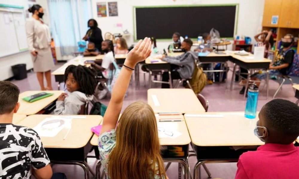 Girl with long blonde hair raises her hand in a classroom; other students sit at desks and a teacher stands near the front.