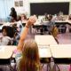 Girl with long blonde hair raises her hand in a classroom; other students sit at desks and a teacher stands near the front.