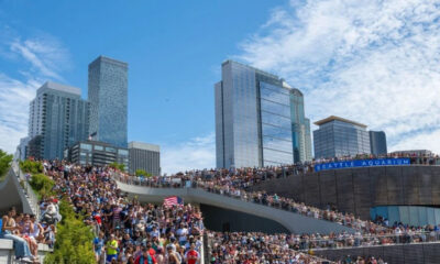 Large crowd on a stepped terrace along the Seattle waterfront, with tall glass skyscrapers in the background and a bright blue sky.