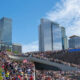 Large crowd on a stepped terrace along the Seattle waterfront, with tall glass skyscrapers in the background and a bright blue sky.