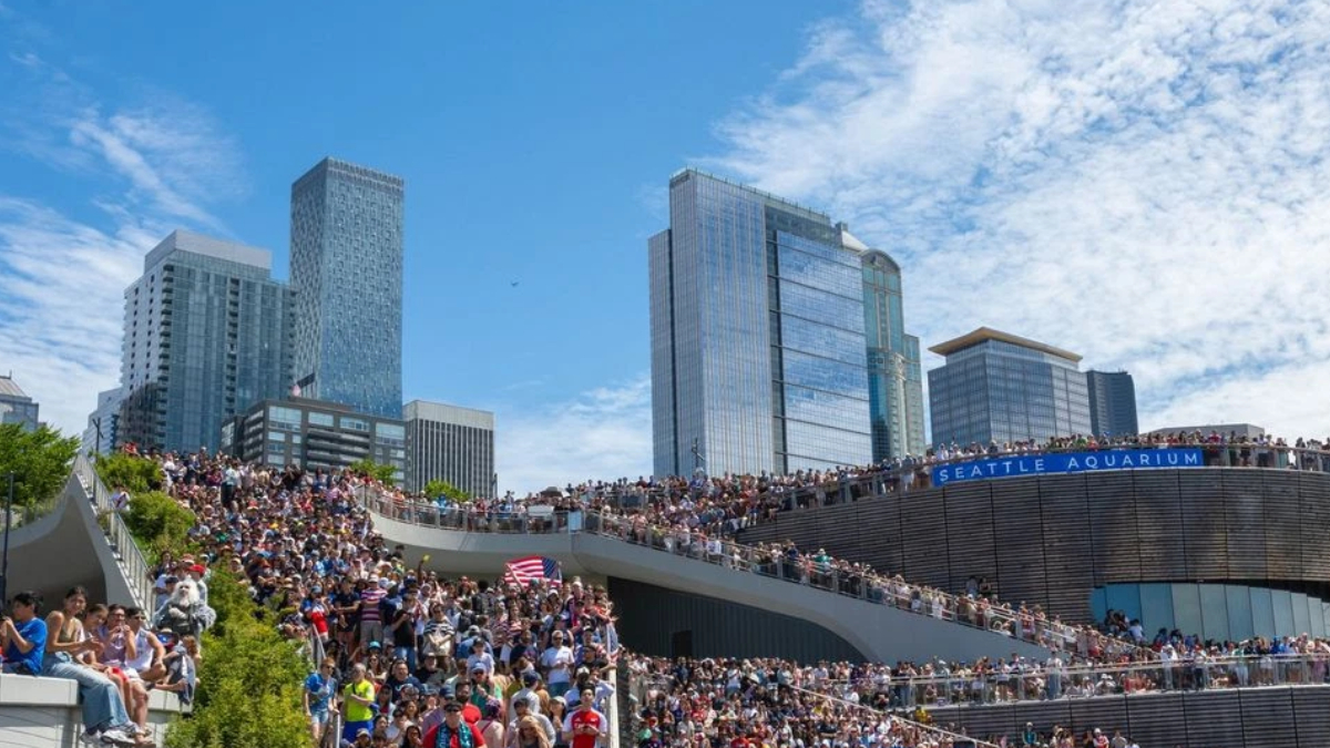 Large crowd on a stepped terrace along the Seattle waterfront, with tall glass skyscrapers in the background and a bright blue sky.