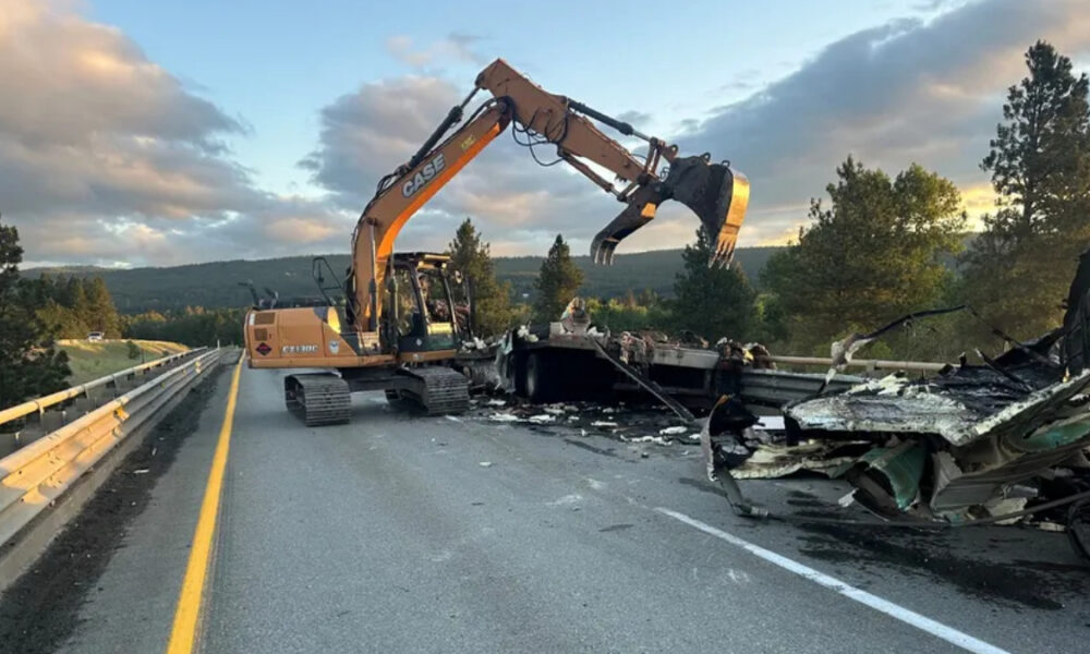 Excavator on a highway clearing wreckage from a crash, with twisted metal and debris across the lanes at sunset. (Informative)