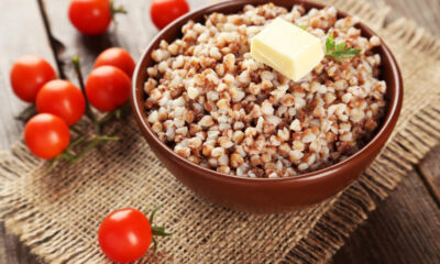Bowl of cooked buckwheat with a pat of butter and a herb sprig, on a rustic wooden table with cherry tomatoes nearby on burlap.