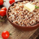 Bowl of cooked buckwheat with a pat of butter and a herb sprig, on a rustic wooden table with cherry tomatoes nearby on burlap.
