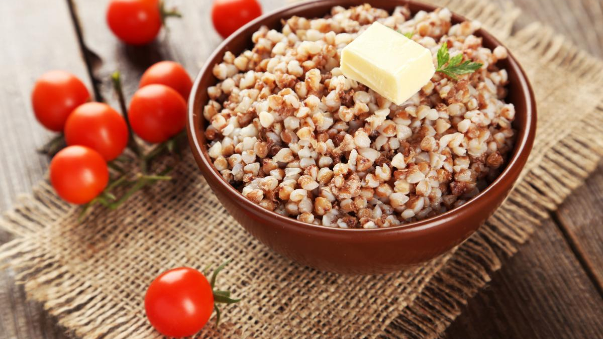 Bowl of cooked buckwheat with a pat of butter and a herb sprig, on a rustic wooden table with cherry tomatoes nearby on burlap.