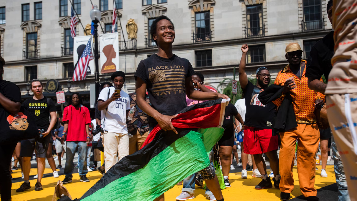Young Black man smiles as he waves a red, black, and green Pan-African flag at a street rally, with diverse protesters and a city building backdrop behind him.