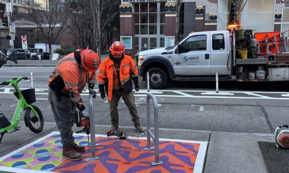 Two construction workers in orange safety gear install a colorful sidewalk mat with bike racks on a city street, SDOT truck in the background.