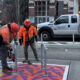 Two construction workers in orange safety gear install a colorful sidewalk mat with bike racks on a city street, SDOT truck in the background.