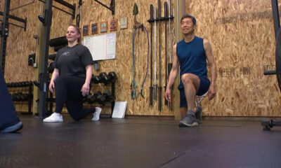 Two people perform kneeling lunges in a gym, with a wooden wall and equipment behind them.