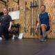 Two people perform kneeling lunges in a gym, with a wooden wall and equipment behind them.
