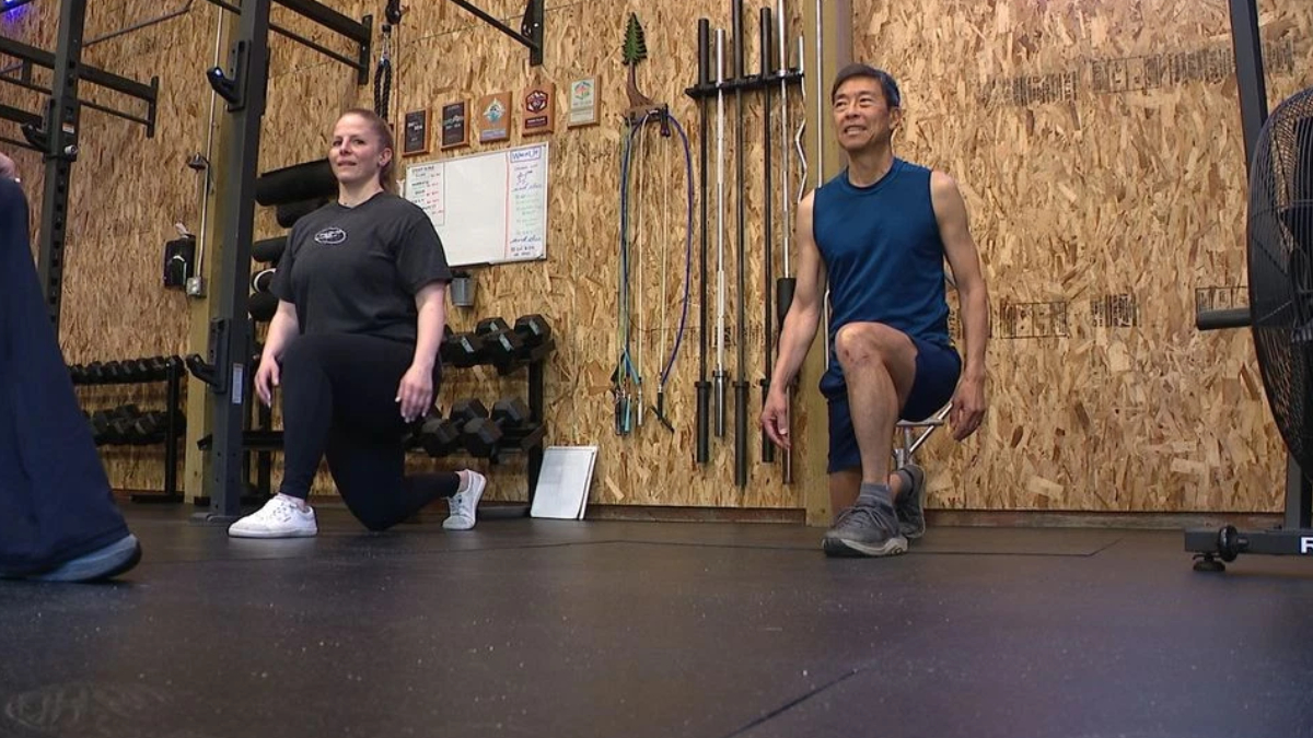 Two people perform kneeling lunges in a gym, with a wooden wall and equipment behind them.
