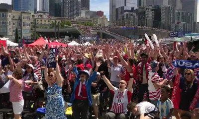 Large crowd of diverse fans cheering with USA flags at an outdoor urban event, tents set up, tall buildings in the background.