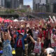 Large crowd of diverse fans cheering with USA flags at an outdoor urban event, tents set up, tall buildings in the background.