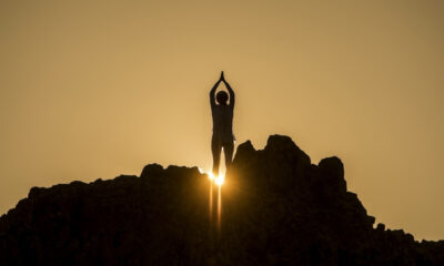 Silhouette of a person standing on jagged rocks at sunset, arms raised overhead, sun bursting behind the silhouette.