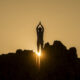 Silhouette of a person standing on jagged rocks at sunset, arms raised overhead, sun bursting behind the silhouette.