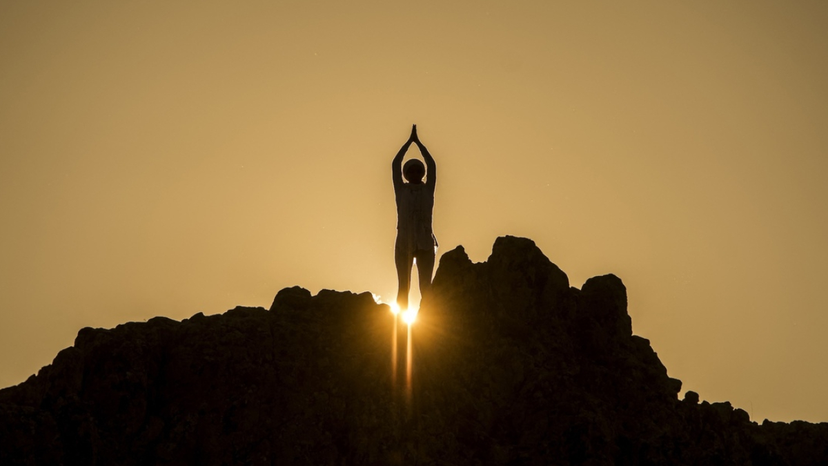 Silhouette of a person standing on jagged rocks at sunset, arms raised overhead, sun bursting behind the silhouette.