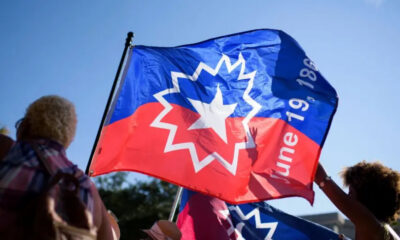 People at a rally waving a large blue and red flag with a white starburst emblem against a clear sky.