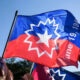 People at a rally waving a large blue and red flag with a white starburst emblem against a clear sky.
