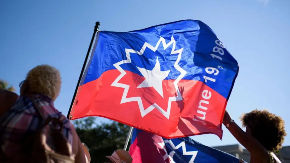 People at a rally waving a large blue and red flag with a white starburst emblem against a clear sky.
