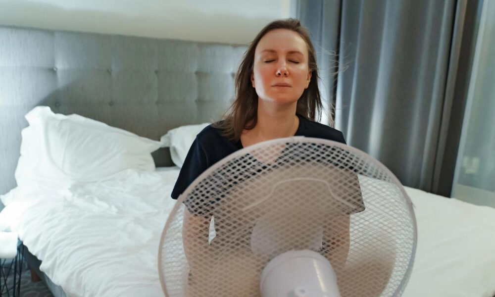 Woman with eyes closed meditating in front of a standing fan in a bedroom with a gray upholstered headboard and white bedding behind her.