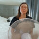 Woman with eyes closed meditating in front of a standing fan in a bedroom with a gray upholstered headboard and white bedding behind her.