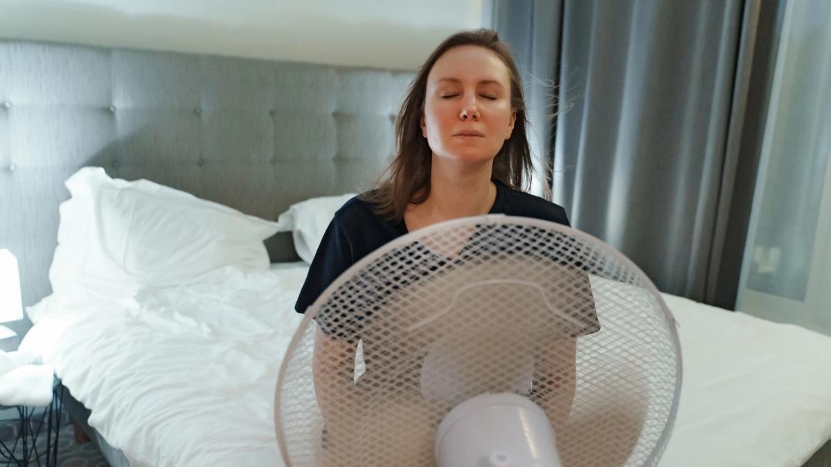 Woman with eyes closed meditating in front of a standing fan in a bedroom with a gray upholstered headboard and white bedding behind her.