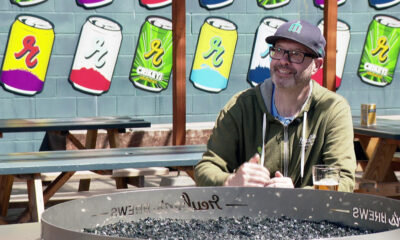Man in a cap and glasses sits at a picnic table outdoors, with a beer glass on the table and a metal fire pit in the foreground, colorful can murals on the wall behind him.