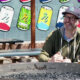 Man in a cap and glasses sits at a picnic table outdoors, with a beer glass on the table and a metal fire pit in the foreground, colorful can murals on the wall behind him.