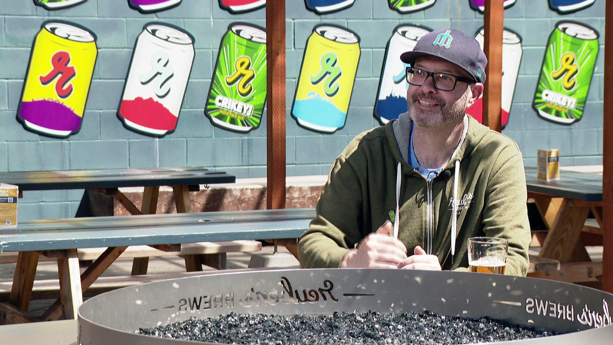 Man in a cap and glasses sits at a picnic table outdoors, with a beer glass on the table and a metal fire pit in the foreground, colorful can murals on the wall behind him.