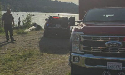 Two law enforcement vehicles parked near a lakeshore while a man stands on a dirt path in the foreground; people are in the water in the background.