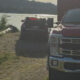 Two law enforcement vehicles parked near a lakeshore while a man stands on a dirt path in the foreground; people are in the water in the background.