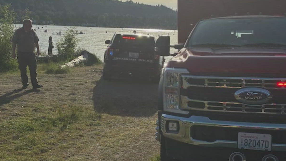 Two law enforcement vehicles parked near a lakeshore while a man stands on a dirt path in the foreground; people are in the water in the background.
