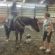 Woman on horseback in an indoor riding arena being greeted by another woman; a small dog sits nearby.